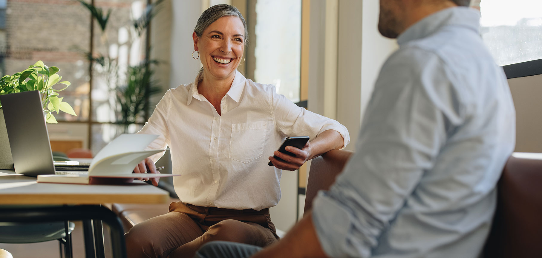 A smiling woman sitting with a phone in hand talking to a man.