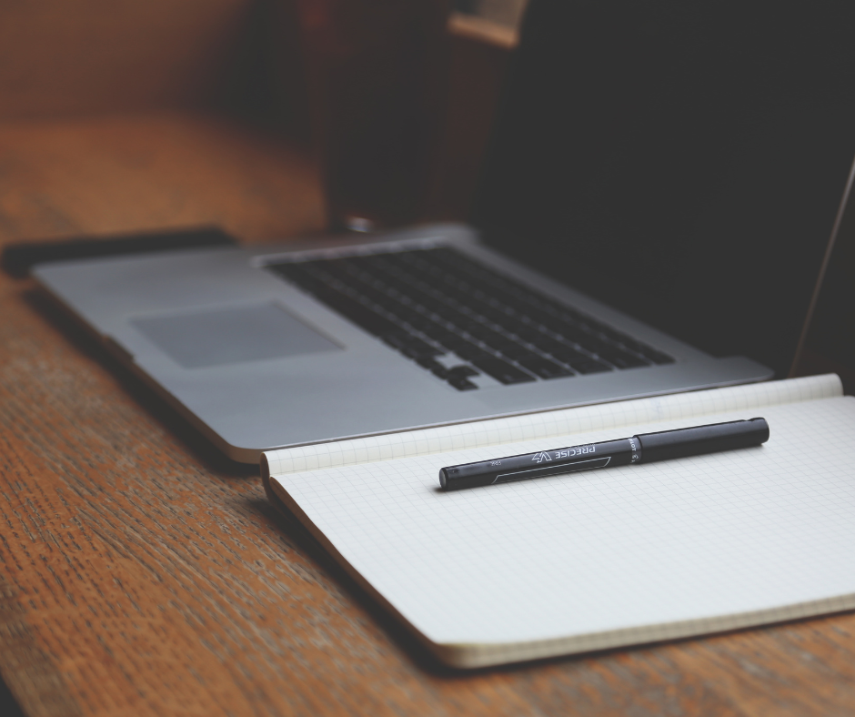 A laptop computer and a notebook with a pen on a table.