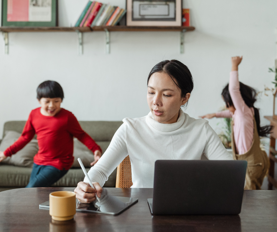 an adult writes on a notepad at a table with a computer. two children move around the room in the background
