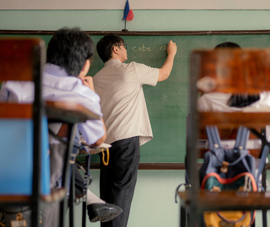 a teacher writes on a chalkboard in the background. students sit in desks facing the chalkboard.