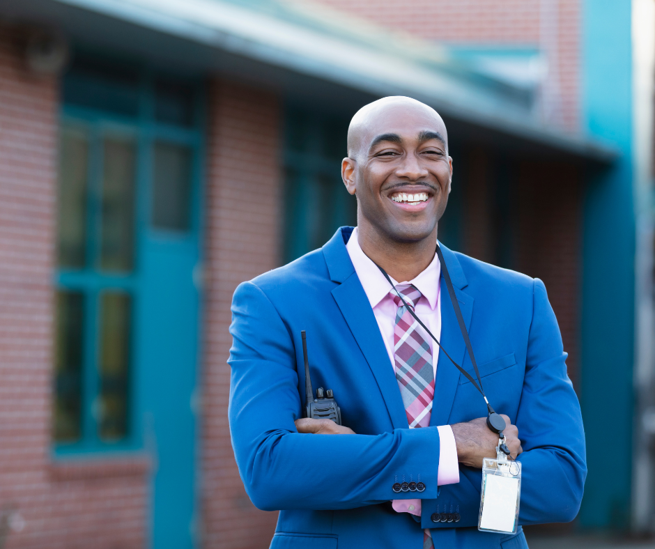 A man stands with arms crossed holding a walkie talkie and wearing a school badge.
