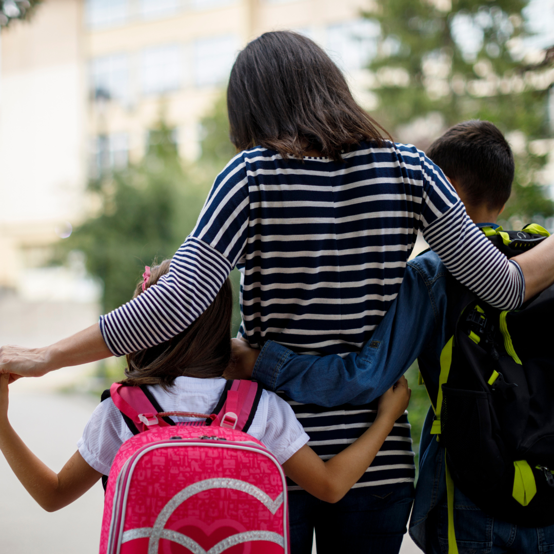 An adult walks with arms around two children wearing backpacks.